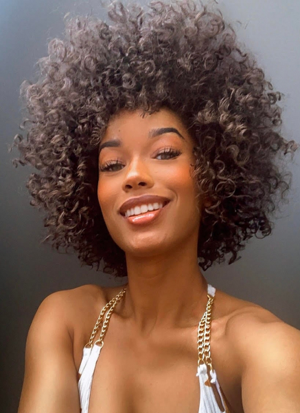 selfie of a woman with curly brown hair wearing a white and gold top