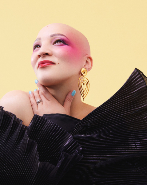 portrait of a bald woman wearing hot pink blush red lipstick blue nails gold drop earrings and a dramatic black top...