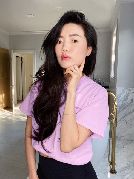 photo of a woman with one hand on her chin long black hair pulled over one shoulder wearing a pink t shirt